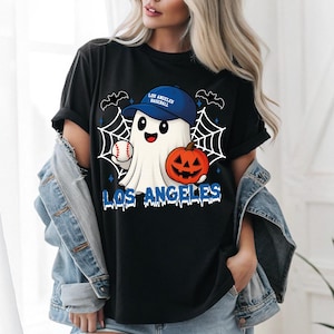 Puede incluir: Camiseta negra con un fantasma de dibujos animados que lleva una gorra de béisbol azul con el texto "Los Angeles Baseball". El fantasma sostiene una pelota de béisbol y una calabaza. Las palabras "Los Angeles" están impresas en azul.