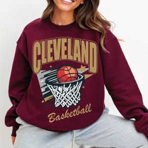 Puede incluir: Una sudadera color granate con un gráfico de un balón de baloncesto entrando en una canasta. El gráfico incluye el texto "CLEVELAND Basketball".