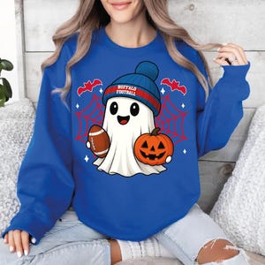 May include: Royal blue sweatshirt featuring a cartoon ghost wearing a blue and red beanie with "BUFFALO FOOTBALL" text. The ghost holds a football and a jack-o'-lantern, with a red spiderweb and bats in the background. A fun, festive design.