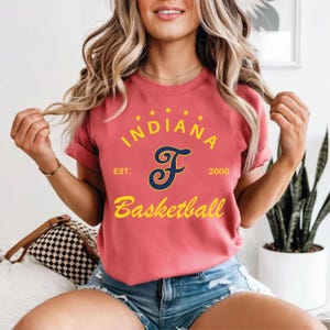 Puede incluir: Una camiseta de color coral con el texto "INDIANA Basketball" en escritura amarilla. La letra "F" es de color azul marino con un remolino decorativo. La camiseta también incluye el texto "EST. 2000" y cinco estrellas amarillas encima de la palabra "INDIANA".