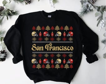Può includere: Felpa nera con un design festivo. Il design include alberi di Natale dorati e rossi, caschi da football e fiocchi di neve. Il testo "San Francisco" è in oro al centro.