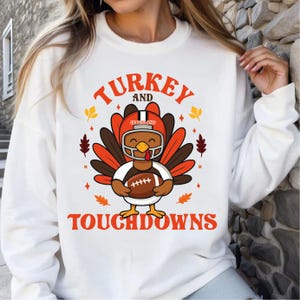 May include: White sweatshirt with a graphic of a turkey wearing a football helmet and holding a football. The text "TURKEY AND TOUCHDOWNS" is in orange. Autumn leaves and a football helmet are also featured in the design.