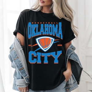 Puede incluir: Camiseta negra con un gráfico de baloncesto azul y naranja y el texto "BASKETBALL OKLAHOMA CITY EST. 2008".