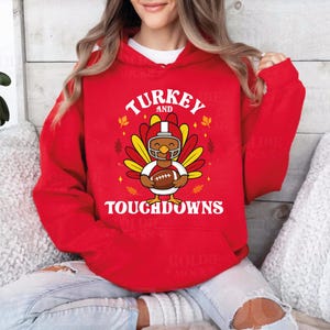 Pode incluir: Um moletom vermelho com as palavras "TURKEY and TOUCHDOWNS" em branco. O gráfico apresenta um peru de desenho animado usando um capacete de futebol e segurando uma bola. O moletom é de cor vermelha lisa.
