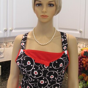 May include: A black and white apron with a red bodice and a floral pattern. The apron has a bow at the waist and a pearl necklace is worn around the neck.