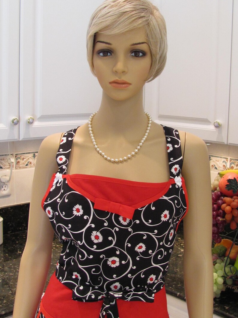 May include: A black and white apron with a red bodice and a floral pattern. The apron has a bow at the waist and a pearl necklace is worn around the neck.