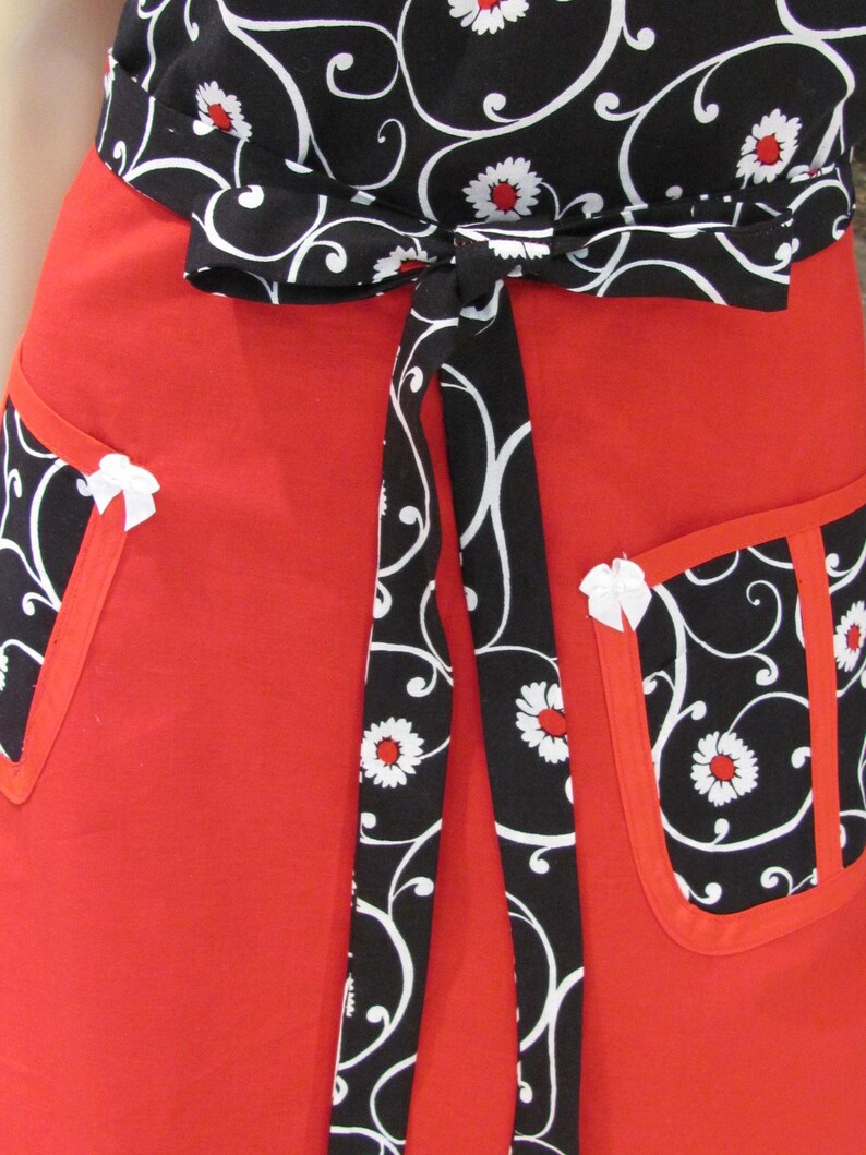 May include: Red apron with black and white floral print. The apron has a tie waist and two pockets.