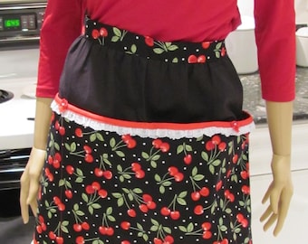 May include: A black apron with a red trim and a white lace border. The apron has a cherry print with green leaves and white polka dots. The apron is being worn by a person with a red shirt.