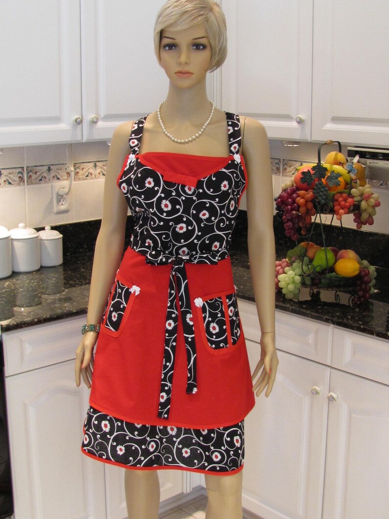 May include: A red and black apron with a floral print and a white trim. The apron has two pockets and a tie at the waist.