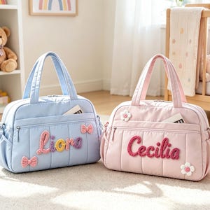 May include: Two quilted duffel bags, one light blue and one light pink, each featuring a zippered front pocket, two top handles, and a detachable shoulder strap. The blue bag displays the name Liora with bow accents, while the pink bag displays the name Cecilia with flower accents.