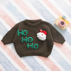 May include: A brown knit Christmas sweater with the words "HO HO HO" in green and a Santa Claus face. The sweater has long sleeves and a round neckline. A wooden toy camera is in the background.