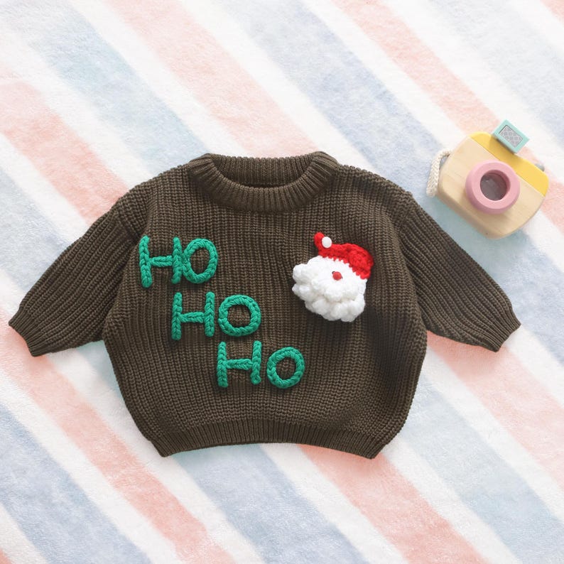 May include: A brown knit Christmas sweater with the words "HO HO HO" in green and a Santa Claus face. The sweater has long sleeves and a round neckline. A wooden toy camera is in the background.