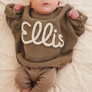 Può includere: Un bambino indossa un maglione marrone a maglia con il nome "Ellis" in corsivo bianco. Il bambino indossa anche pantaloni marroni abbinati. Il maglione ha una trama a coste e uno scollo tondo. Il bambino è sdraiato su un lenzuolo color crema.