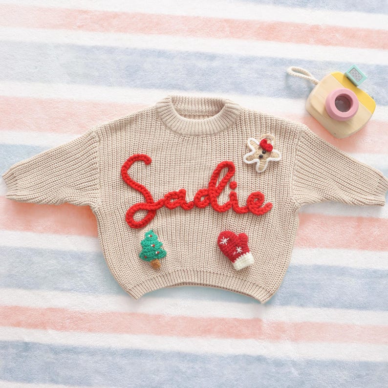 May include: A beige knit sweater with the name "Sadie" in red script. The sweater is decorated with a gingerbread man, Christmas tree, and mitten appliqu&eacute;s. A wooden toy camera is in the background.
