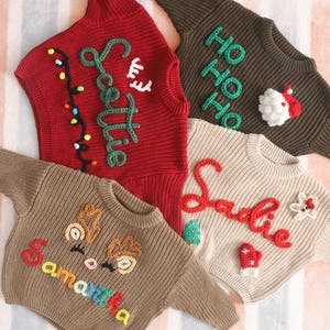 May include: Four knitted Christmas sweaters in red, brown, and beige. Each sweater features a name or holiday greeting in colorful yarn. Decorations include a string of lights, reindeer, and a Santa Claus face.