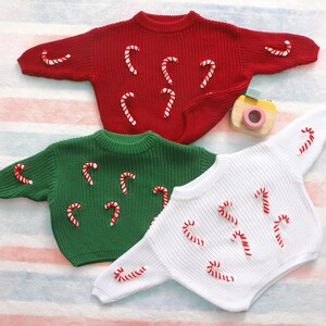 May include: Three knitted sweaters in red, green, and white, each adorned with candy cane appliqu&eacute;s. The sweaters have a round neckline and long sleeves. A small toy camera is visible in the background.