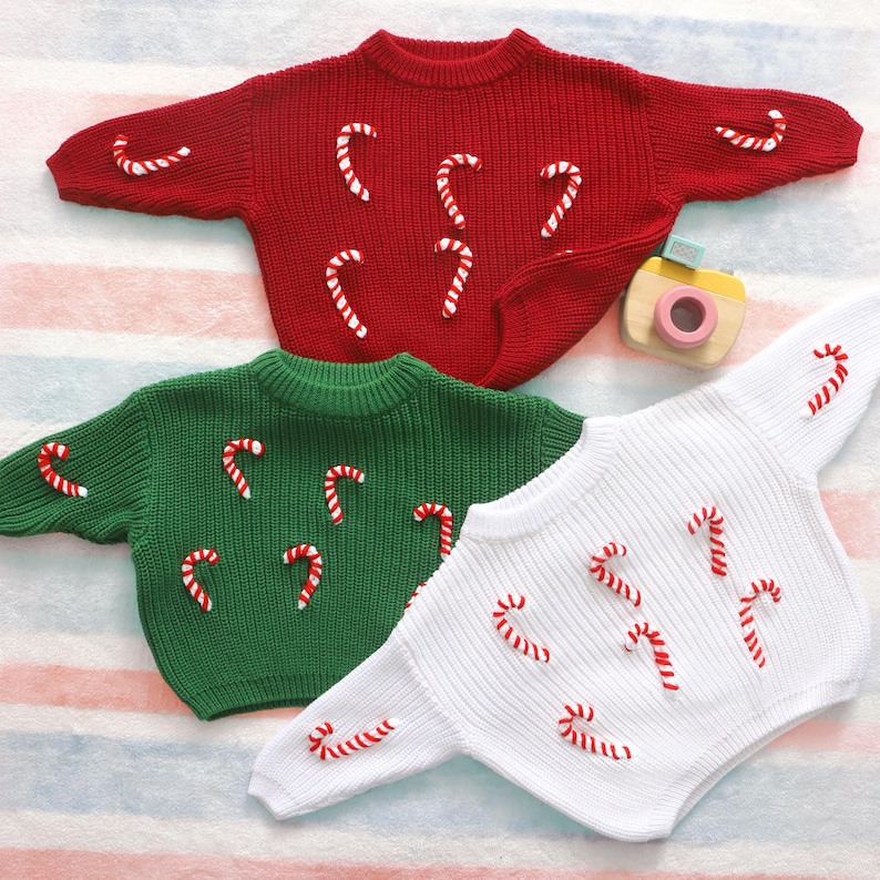 May include: Three knitted sweaters in red, green, and white, each adorned with candy cane appliqu&eacute;s. The sweaters have a round neckline and long sleeves. A small toy camera is visible in the background.