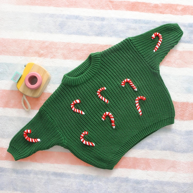 May include: A green knit sweater decorated with red and white candy cane appliqu&eacute;s. The sweater has a round neckline and long sleeves. A small wooden toy camera with a pink lens sits to the left of the sweater.