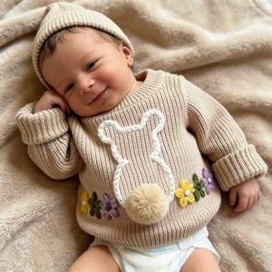 May include: A baby in a beige knitted sweater and beanie. The sweater has a white bunny design, a pom-pom tail, and embroidered flowers in yellow, green, and purple. The baby is smiling, lying on a beige blanket.