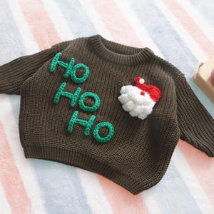 May include: A brown knitted sweater with the words "HO HO HO" in green, and a Santa Claus applique. The sweater is made of a soft, textured material, perfect for the holiday season. The Santa applique features a white beard and a red hat.