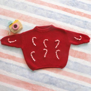 May include: A red knit sweater decorated with white and red candy cane appliqu&eacute;s. The sweater has a crew neck and long sleeves. A wooden toy camera is in the upper left corner. The sweater is on a striped background.