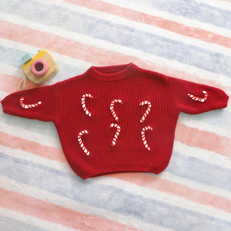 May include: A red knit sweater decorated with white and red candy cane appliqu&eacute;s. The sweater has a crew neck and long sleeves. A wooden toy camera is in the upper left corner. The sweater is on a striped background.