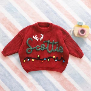 May include: A red knit sweater with the name "Scottie" in green script and white antlers. The sweater has a string of colorful lights embroidered across the bottom. A wooden toy camera is in the background.