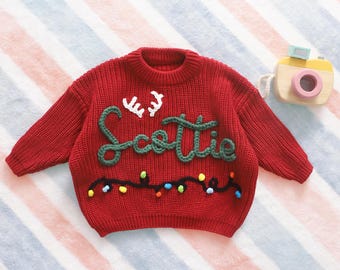 May include: A red knit sweater with the name "Scottie" in green script and white antlers. The sweater has a string of colorful lights embroidered across the bottom. A wooden toy camera is in the background.