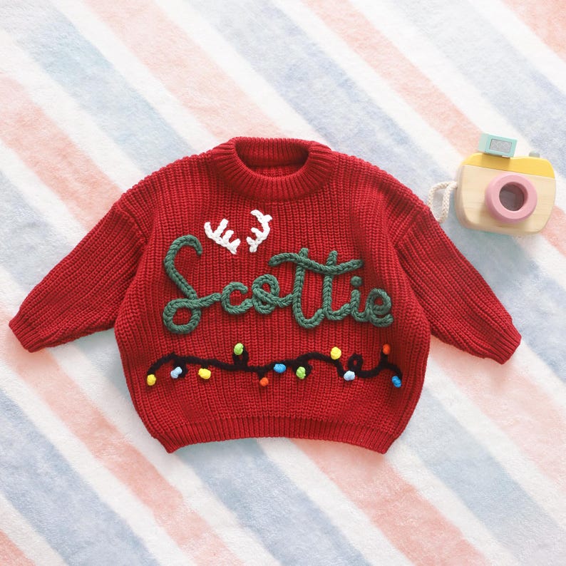May include: A red knit sweater with the name "Scottie" in green script and white antlers. The sweater has a string of colorful lights embroidered across the bottom. A wooden toy camera is in the background.