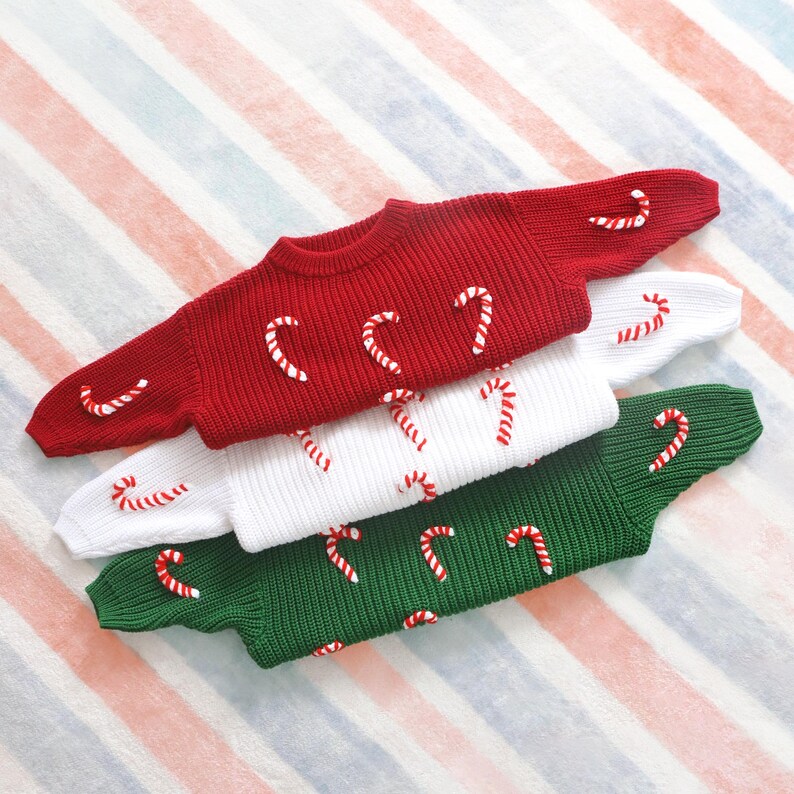 May include: Three knitted sweaters in red, white, and green, each adorned with candy cane appliqu&eacute;s. The sweaters are stacked, showcasing the festive holiday theme. The background features a striped pattern in red, white, and blue.