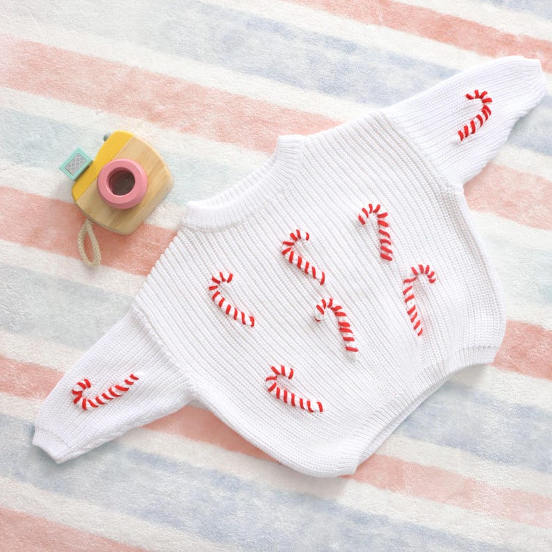 May include: White knit sweater with red candy cane embroidery. The sweater has a round neck and long sleeves. A wooden toy camera with a pink lens is next to the sweater on a striped background.