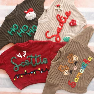 May include: A collection of Christmas-themed sweaters in various colors, including brown, red, and beige. Each sweater features unique embroidered designs such as "Ho Ho Ho", "Scottie", "Joy", and "Samantha", along with festive embellishments like Santa, reindeer, and lights.