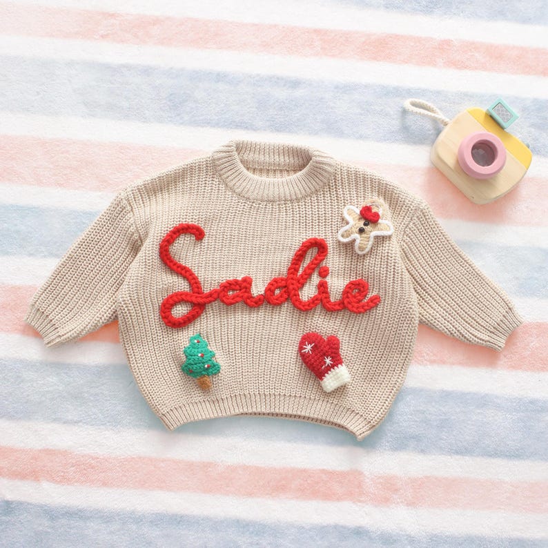 May include: Beige knit sweater with the name "Sadie" in red script. The sweater features a Christmas tree, a gingerbread man, and a mitten applique. A toy camera rests on the striped background.