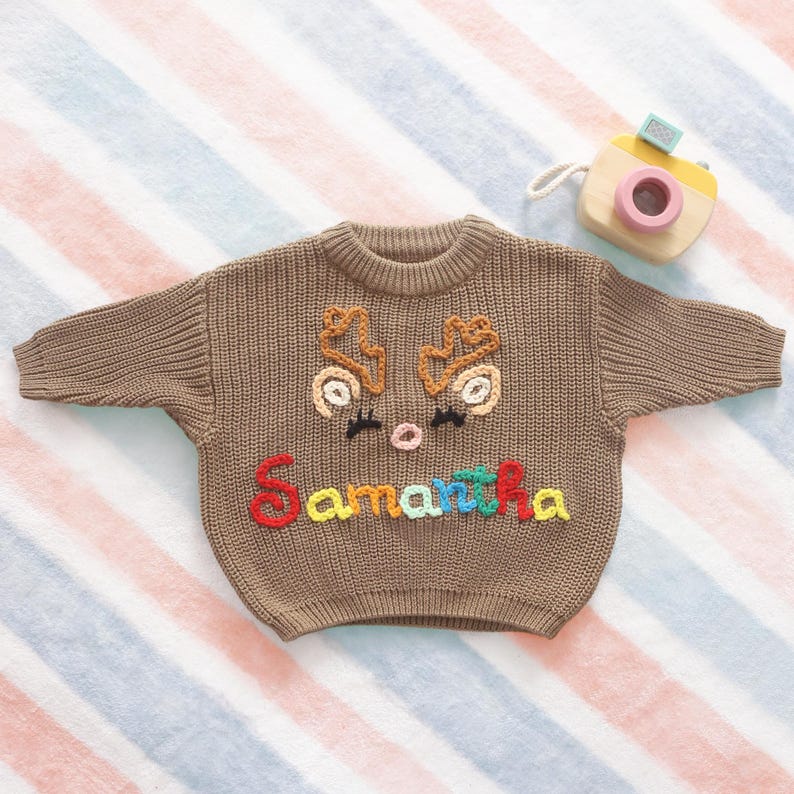 May include: A brown knit sweater with a reindeer face design and the name "Samantha" in colorful embroidery. The sweater has a round neck and long sleeves. A wooden toy camera is in the background.