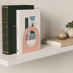 May include: A pink metal bookend with a rounded arch design holds two books on a white shelf. One book is white with a geometric design, and the other is dark green. The bookend has the text "IDGE THER".