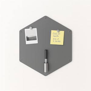 May include: A gray, hexagonal magnetic memo board with a silver pen holder. It has a small photo and a yellow sticky note that reads "Call Dr. Smith 9/11 at 4:30 PM" attached with magnets. The board has a minimalist design.