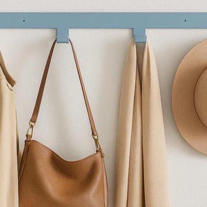 May include: A light blue wall-mounted coat rack with three hooks, holding a tan trench coat, a brown leather handbag, and a tan scarf. A tan fedora hat hangs on the right. The items are against a white wall.