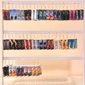 May include: A white metal earring display stand with three tiers. Each tier holds multiple pairs of book-themed earrings. The books include titles like "The Hunger Games", "Twilight", "Percy Jackson", and "Harry Potter".