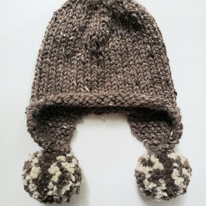 May include: A hand-knitted brown hat with ear flaps and two pom-poms. The hat is made of thick, textured yarn with flecks of white and black. The pom-poms are a mix of brown and white yarn.