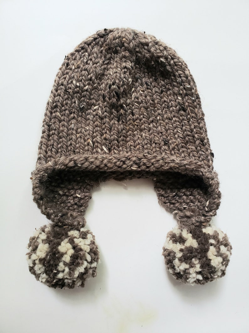 May include: A hand-knitted brown hat with ear flaps and two pom-poms. The hat is made of thick, textured yarn with flecks of white and black. The pom-poms are a mix of brown and white yarn.