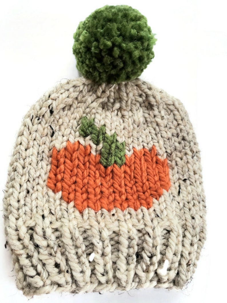 May include: A hand-knitted beanie hat with a pumpkin design. The hat is beige with flecks of black and features an orange pumpkin applique with green leaves. A large green pom-pom tops the hat.