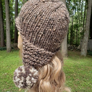 May include: A brown knit hat with a brim and ear flaps, featuring a large pom-pom. The hat is made of a chunky, textured yarn with flecks of white and brown. The hat is designed to keep the wearer warm.