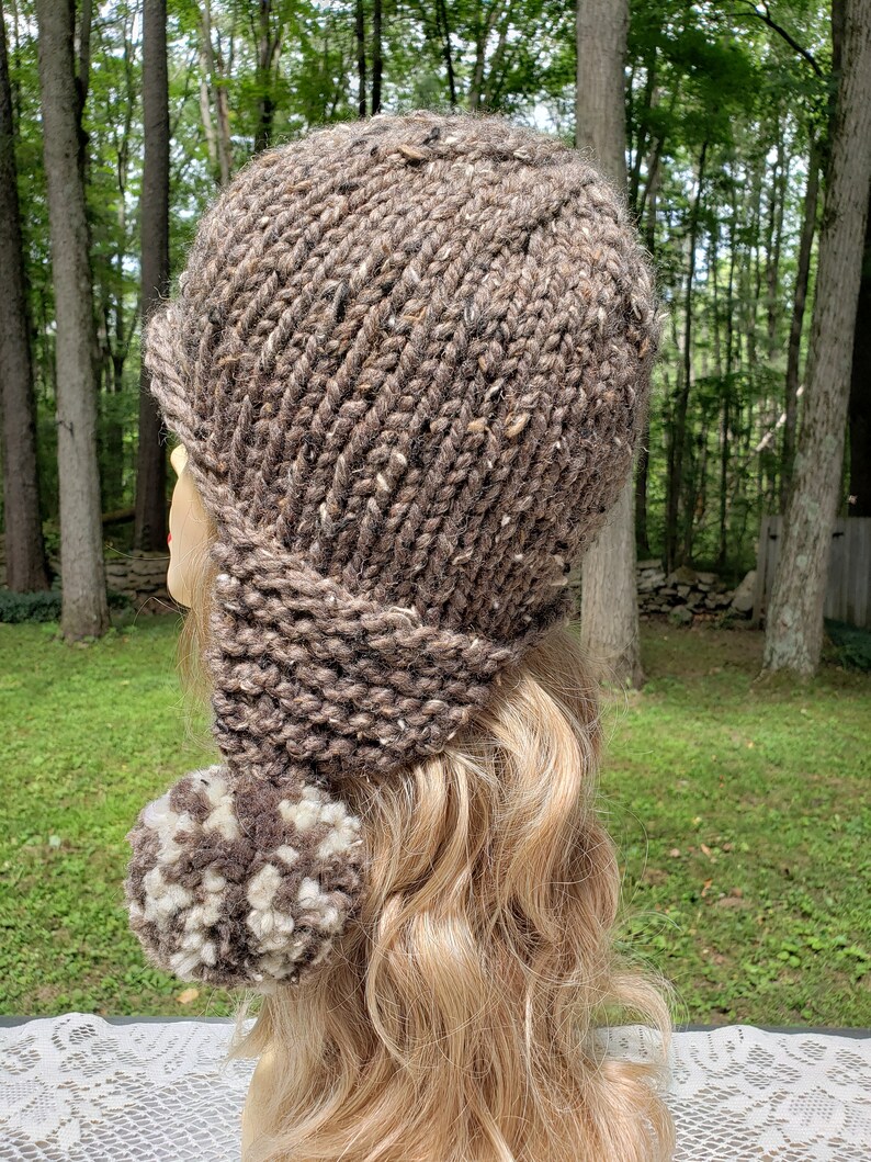 May include: A brown knit hat with a brim and ear flaps, featuring a large pom-pom. The hat is made of a chunky, textured yarn with flecks of white and brown. The hat is designed to keep the wearer warm.