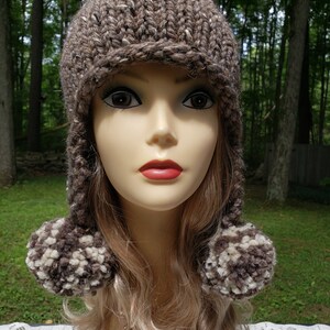 May include: A hand-knitted brown hat with ear flaps and two pom-poms. The hat is made of a thick, textured yarn with flecks of white and brown. The pom-poms are a mix of brown and cream-colored yarn. The hat is displayed on a mannequin head.