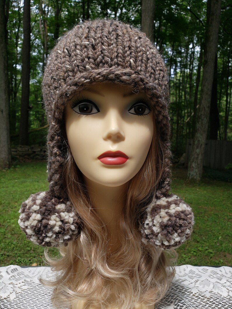 May include: A hand-knitted brown hat with ear flaps and two pom-poms. The hat is made of a thick, textured yarn with flecks of white and brown. The pom-poms are a mix of brown and cream-colored yarn. The hat is displayed on a mannequin head.