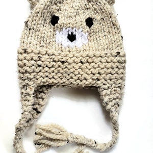 May include: A beige knitted hat with bear ears and a black and white bear face design. The hat has long ties with tassels.