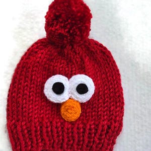 May include: A red knit hat with a large pom-pom on top. The hat features white and black eyes and an orange nose, resembling a popular children's character. The hat is made of a thick, textured yarn.