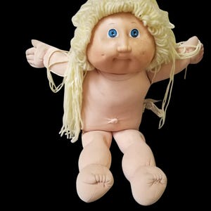 Op de afbeelding: Een vintage Cabbage Patch Kid pop met een zacht, vleeskleurig lichaam en lichtblond garen haar. De pop heeft blauwe ogen en poseert met uitgestrekte armen. De pop is een klassiek verzamelobject.