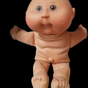 Puede incluir: Una muñeca Cabbage Patch Kid vintage con cabeza calva, cara redonda y ojos pintados. La muñeca tiene un cuerpo suave color carne con los brazos extendidos y pies detallados. La muñeca está sobre un fondo negro.