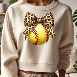 Pode incluir: Uma camisa de moletom cor creme com uma bola de softball e um laço de estampa de leopardo. A bola de softball é amarela com costuras vermelhas. O laço está amarrado em volta da bola de softball.